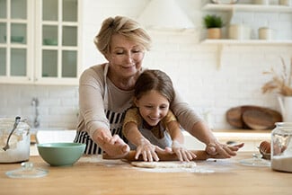 Grandmother reaching over granddaughter to help her roll out cookie dough