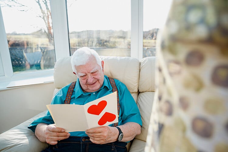 Elderly White haired man sitting in a comfy chair reading a card hand made by a child